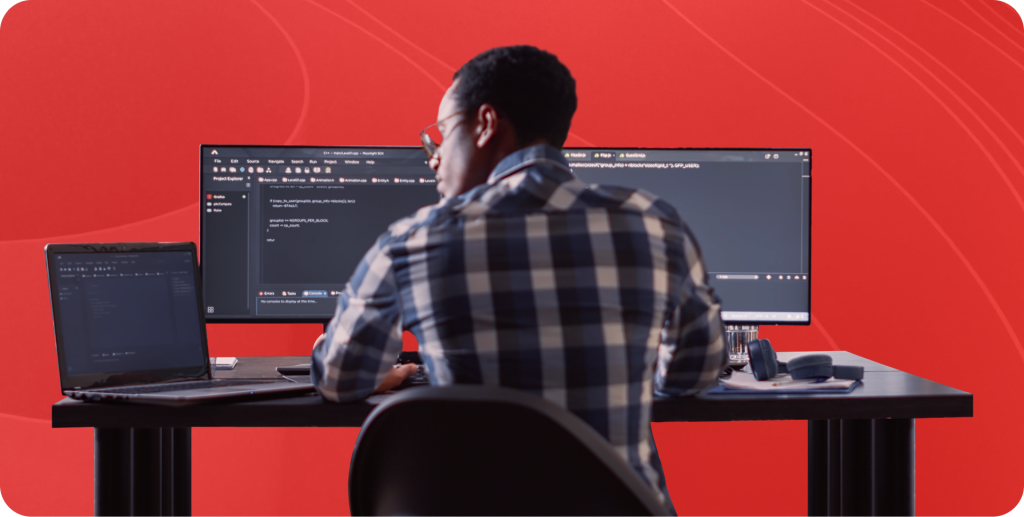 A DevOps engineer sits at a desk in front of a laptop and 2 monitors coding.
