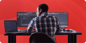 A DevOps engineer sits at a desk in front of a laptop and 2 monitors coding.