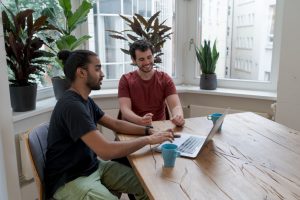 Two developers discuss a project in a bright office with indoor plants on the windowsill. There is a laptop and two blue mugs on the table in front of them.