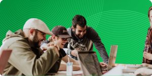 3 coworkers are seen grouped together around laptops at a table. Part of a woman is seen on the right side.