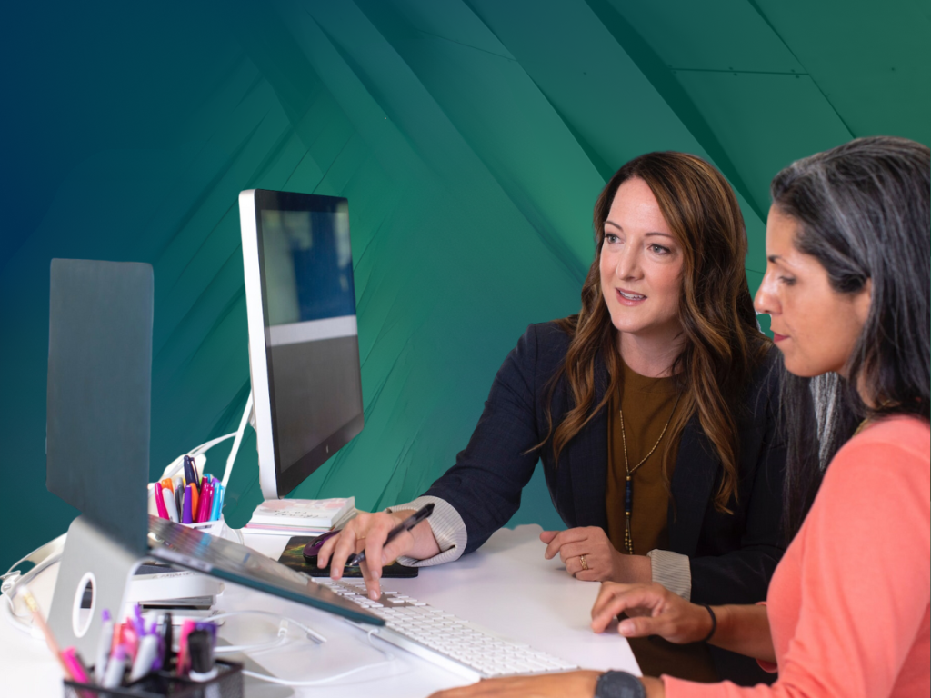 Two coworkers work together at a desk looking at a monitor and computer.