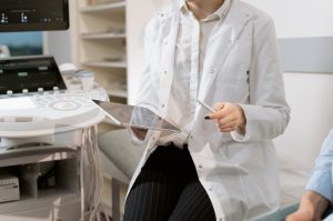A doctor sits between a patient and an ultrasound, holding a tablet that adheres to HIPAA data storage requirements.