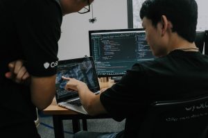 Two coworkers in black T-shirts, one sitting and one standing, in front of a laptop and a computer monitor on a desk. The sitting worker points at code on the laptop screen.