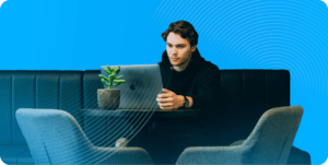 A man in a dark hoodie opens his laptop while sitting on the edge of a clean, black leather couch. The table he rests his laptop on has a small potted plant and two chairs placed around it.